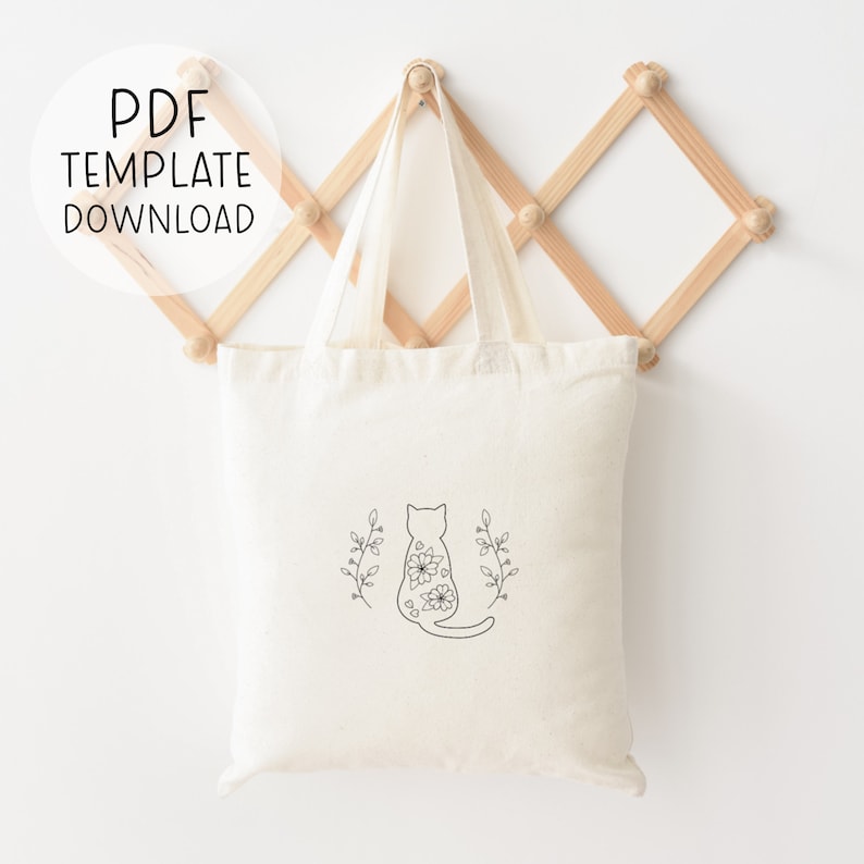 May include: A white canvas tote bag with a black line drawing of a cat with flowers on its back. The cat is sitting with its tail curled around its legs. The cat is surrounded by two sprigs of leaves.
