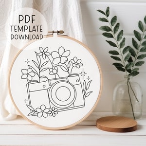 May include: A black and white line drawing of a camera with flowers around it. The image is in a white embroidery hoop.