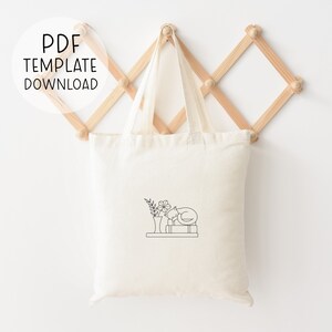 Peut inclure: Un sac en toile blanc avec un dessin au trait noir d'un chat dormant sur une pile de livres avec un vase de fleurs &agrave; c&ocirc;t&eacute;. Le sac en toile est suspendu &agrave; un support en bois.