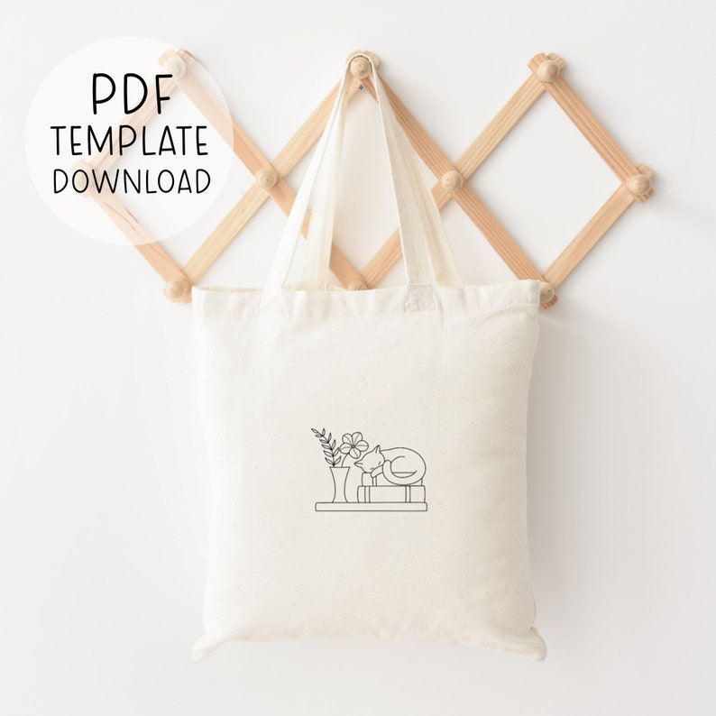 Peut inclure: Un sac en toile blanc avec un dessin au trait noir d'un chat dormant sur une pile de livres avec un vase de fleurs &agrave; c&ocirc;t&eacute;. Le sac en toile est suspendu &agrave; un support en bois.