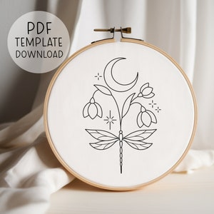 Puede incluir: Un patrón de bordado en blanco y negro de una libélula con una luna creciente y flores. El patrón está en una tela blanca en un aro de bordado de madera.
