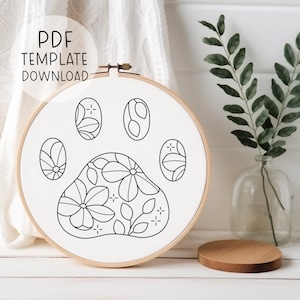 May include: A black and white line drawing of a paw print with floral designs inside. The paw print is in a wooden embroidery hoop.