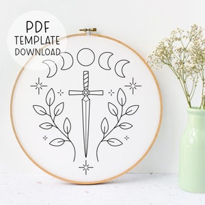 May include: A black and white line drawing of a sword with a cross guard, surrounded by four crescent moons, two leafy branches, and four stars. The design is on a white background inside a wooden embroidery hoop.