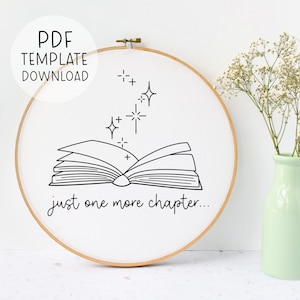 Puede incluir: Un aro de bordado blanco con un dibujo de línea negra de un libro abierto con estrellas arriba. El texto "just one more chapter..." está debajo del libro.