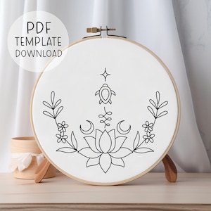 Puede incluir: Un patrón de bordado en blanco y negro en un aro blanco. El patrón presenta una flor de loto, dos lunas crecientes, una tortuga, una estrella y dos ramas de hojas con flores.