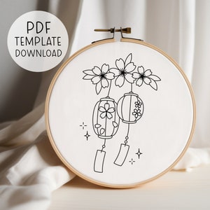 May include: An embroidery hoop with a white fabric featuring a black line drawing of cherry blossoms and Japanese lanterns. The text "PDF TEMPLATE DOWNLOAD" is in a white circle in the upper left corner. The hoop is made of wood.