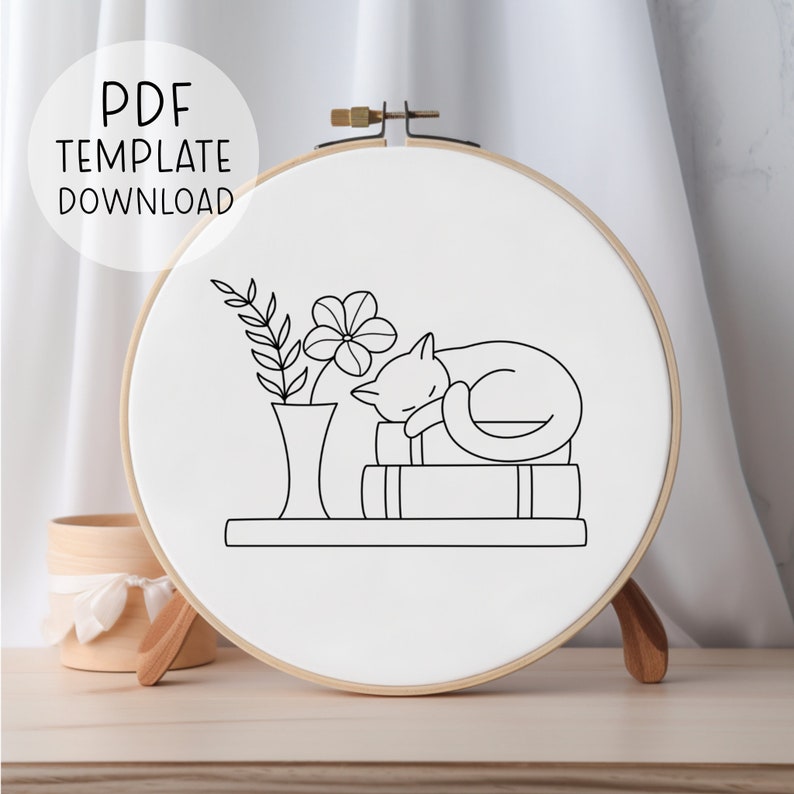 Peut inclure: Dessin au trait noir et blanc d'un chat dormant sur une pile de livres avec un vase et une fleur &agrave; c&ocirc;t&eacute;. Le dessin est sur fond blanc et se trouve dans un cercle de broderie rond.
