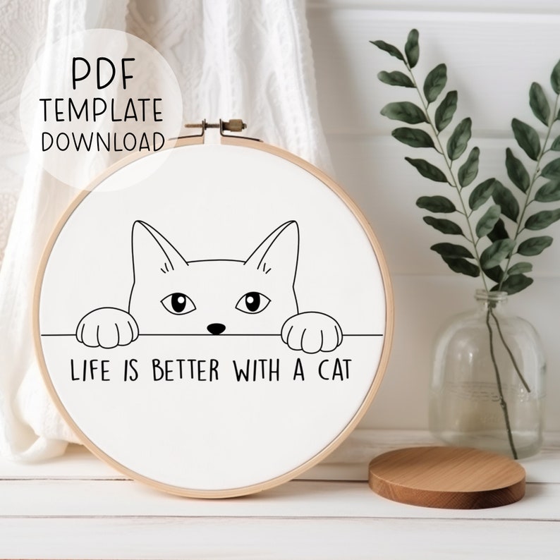 Puede incluir: Un aro de bordado blanco con un dibujo de l&iacute;nea negra de un gato mirando por encima. El texto "Life is better with a cat" est&aacute; impreso debajo del gato.