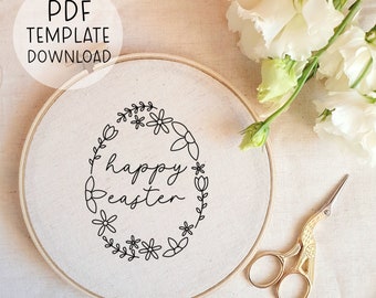 Op de afbeelding: Een witte borduurring met een zwarte omtrek van een bloemenkrans en de tekst "happy easter" erin. De ring ligt op een wit stofoppervlak met witte bloemen en gouden borduurscharen.