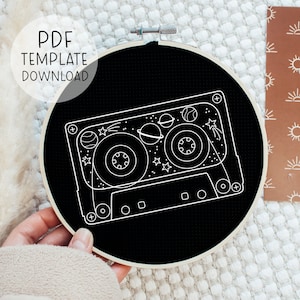 May include: A black and white embroidery hoop with a white outline of a cassette tape with planets and stars inside. The text "PDF TEMPLATE DOWNLOAD" is in the upper left corner of the image.