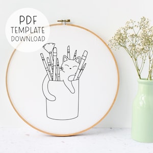 Puede incluir: Dibujo en blanco y negro de una línea de un gato dentro de una taza con pinceles que sobresalen de la parte superior. El gato está durmiendo y tiene una expresión feliz. La imagen está en un aro de bordado redondo.