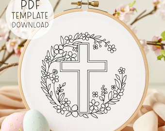 May include: A black and white embroidery design of a cross surrounded by a floral wreath. The design is on a white fabric stretched over a wooden embroidery hoop. The hoop is sitting on a table with two Easter eggs.