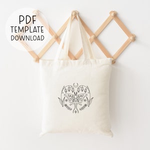 May include: A white tote bag with a black line drawing of a moth, flowers, and a crescent moon. The tote bag is hanging on a wooden rack.