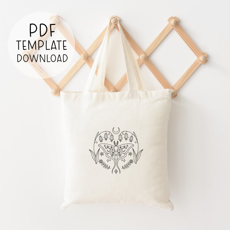 May include: A white tote bag with a black line drawing of a moth, flowers, and a crescent moon. The tote bag is hanging on a wooden rack.