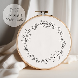 May include: A white embroidery hoop with a black floral wreath design. The design features simple leaves and small flowers.