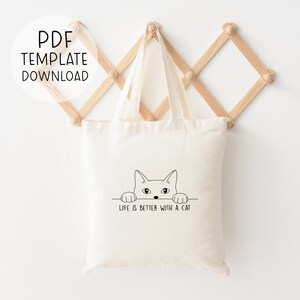 Puede incluir: Una bolsa de tela blanca con un dibujo de l&iacute;nea negra de un gato mirando por encima. El texto "Life is better with a cat" est&aacute; impreso debajo del gato.