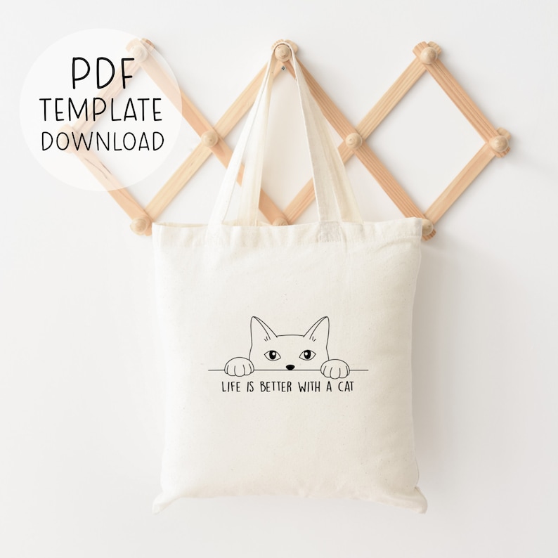 Puede incluir: Una bolsa de tela blanca con un dibujo de l&iacute;nea negra de un gato mirando por encima. El texto "Life is better with a cat" est&aacute; impreso debajo del gato.