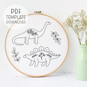 May include: A black and white embroidery pattern featuring two dinosaurs with floral details. The pattern is displayed on a white embroidery hoop.