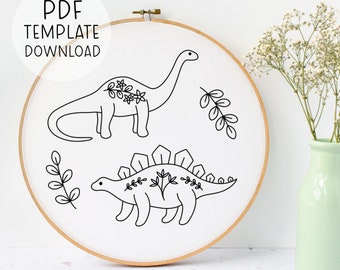 May include: A wooden embroidery hoop containing white fabric with black line art of a long-necked dinosaur and a stegosaurus, both decorated with floral patterns, alongside two leafy sprigs. The text PDF TEMPLATE DOWNLOAD is visible.