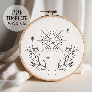 May include: A black and white embroidery hoop with a moon and stars design. The design includes two floral sprigs with small flowers and leaves. The hoop is on a white fabric background.
