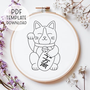 Könnte beinhalten: Eine Schwarzweiß-Zeichnung eines Maneki-Neko, einer japanischen Glückskatze, mit der rechten Pfote erhoben in einer winkenden Geste. Die Katze hält ein rundes Objekt mit dem japanischen Schriftzeichen für "Glück" darauf. Die Zeichnung befindet sich auf einem weißen Hintergrund in einem hölzernen Stickrahmen.