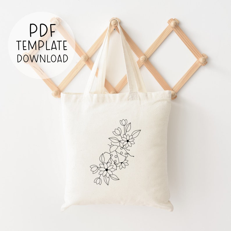 Op de afbeelding: Een witte canvas tote bag met een zwarte lijntekening van een lieveheersbeestje en bloemen aan de voorkant. De tas hangt aan een houten rek.