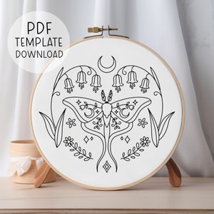 Puede incluir: Un dibujo en blanco y negro de una polilla con una luna creciente en sus alas, rodeado de flores y hojas. El diseño está en un aro de bordado blanco.