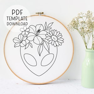 Peut inclure: Motif de broderie en ligne noire et blanche d'un extraterrestre portant une couronne de fleurs. La couronne comprend un hibiscus, une marguerite et d'autres éléments floraux.