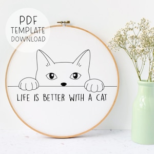 Puede incluir: Una ilustraci&oacute;n en blanco y negro de un gato mirando por encima de una superficie con el texto "Life is better with a cat". La ilustraci&oacute;n est&aacute; sobre un fondo blanco dentro de un aro de bordado de madera.