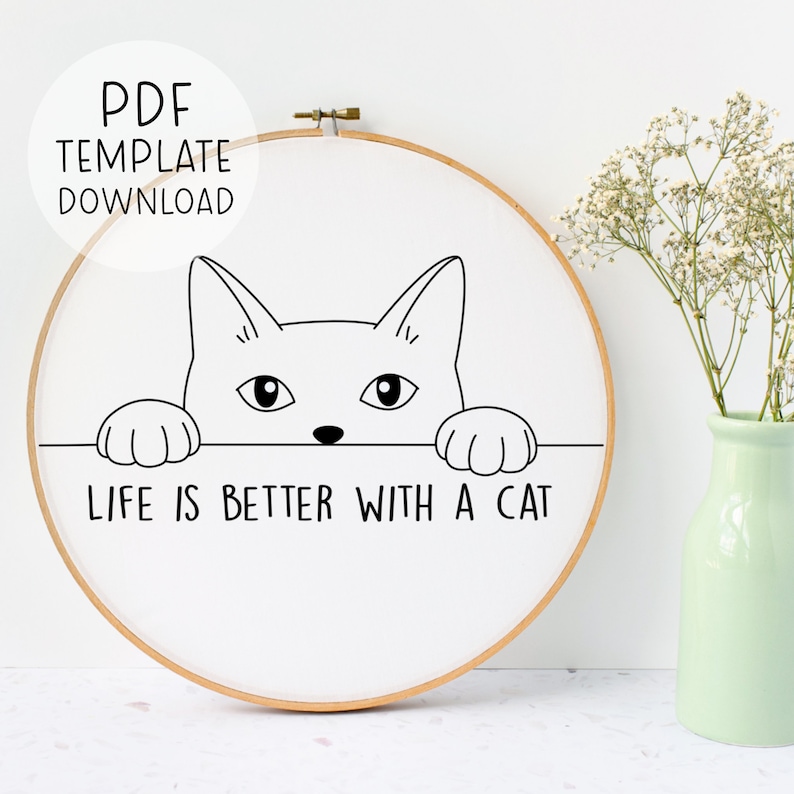 Puede incluir: Una ilustraci&oacute;n en blanco y negro de un gato mirando por encima de una superficie con el texto "Life is better with a cat". La ilustraci&oacute;n est&aacute; sobre un fondo blanco dentro de un aro de bordado de madera.