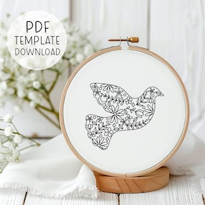 Puede incluir: Un dibujo en blanco y negro de una paloma con un patrón floral. La paloma está en un aro de bordado blanco con un marco de madera.