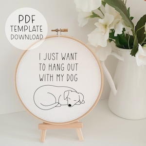 Peut inclure: Un cercle de broderie blanc avec le texte "I just want to hang out with my dog" et un dessin au trait noir d'un chien endormi. Le cercle est sur un chevalet en bois.