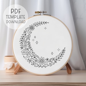 May include: A black and white embroidery pattern of a crescent moon with floral details, stars, and leaves. The pattern is displayed on a white embroidery hoop.