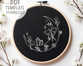 Può includere: Un cerchio da ricamo nero con un design a forma di luna crescente bianca. La luna è decorata con una falena, cristalli, funghi e fiori.