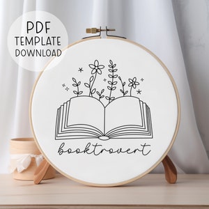 May include: A black and white embroidery pattern of an open book with flowers growing out of it. The text "booktrovert" is below the book.