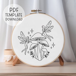 May include: A black and white line drawing embroidery pattern of two fairies with wings and a mushroom with three caps. The pattern is on a white background and is in a round embroidery hoop.