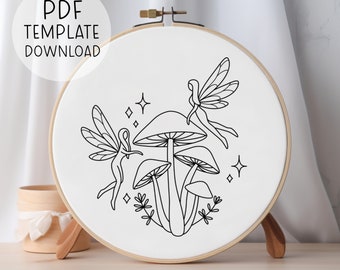 May include: A black and white line drawing embroidery pattern of two fairies with wings and a mushroom with three caps. The pattern is on a white background and is in a round embroidery hoop.