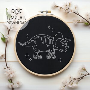 Könnte beinhalten: Ein schwarzer Stickrahmen mit einer weißen Umrandung eines Triceratops-Skeletts. Das Skelett ist von kleinen weißen Sternen und Pluszeichen umgeben.