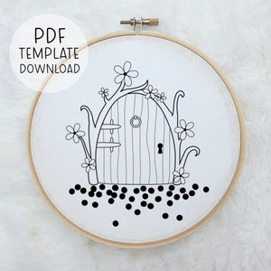 Pode incluir: Um desenho a preto e branco de uma porta de fada caprichosa com uma fechadura, rodeada de flores e videiras, sobre um fundo branco. A porta est&aacute; sobre um tecido branco esticado sobre um aro de bordado de madeira.