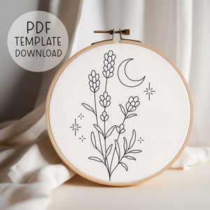 Puede incluir: Patrón de bordado de línea en blanco y negro de flores de lavanda, una luna creciente y estrellas. El patrón está en una tela blanca en un aro de bordado de madera.