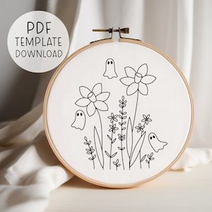 May include: A black and white embroidery pattern featuring three ghosts and daffodils with leaves and stems. The pattern is on a white fabric stretched over a wooden embroidery hoop.