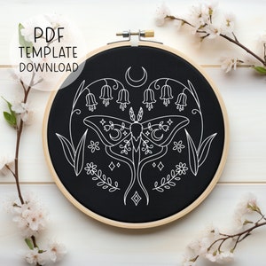 May include: A black embroidery hoop with a white line drawing of a moth, flowers, and crescent moons. The design is centered in the hoop and has a minimalist aesthetic.