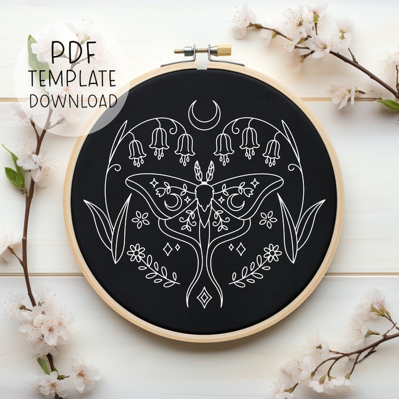 May include: A black embroidery hoop with a white line drawing of a moth, flowers, and crescent moons. The design is centered in the hoop and has a minimalist aesthetic.