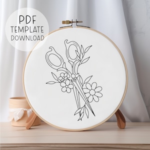 May include: A black and white embroidery pattern of a pair of scissors with floral accents on a white embroidery hoop.