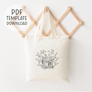May include: A white canvas tote bag with a black line drawing of mushrooms and flowers in a circle. The tote bag has black handles and is hanging on a wooden rack.