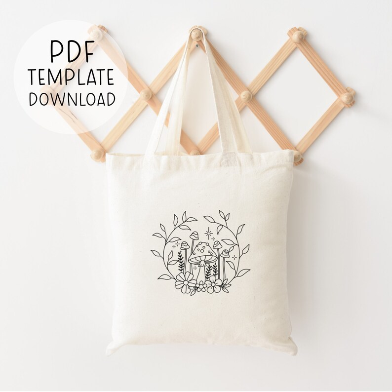 May include: A white canvas tote bag with a black line drawing of mushrooms and flowers in a circle. The tote bag has black handles and is hanging on a wooden rack.