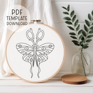 May include: A black and white line drawing of a butterfly with scissors for wings, displayed in a wooden embroidery hoop. The hoop is on a white surface.