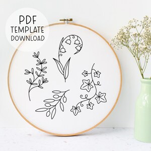 Pode incluir: Um padr&atilde;o de bordado preto e branco com quatro desenhos florais diferentes: l&iacute;rios do vale, hera, ramos de oliveira e um galho simples. O padr&atilde;o &eacute; exibido em um tecido branco esticado sobre um aro de bordado de madeira.