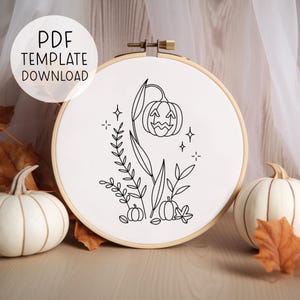 以下が含まれることがあります： 笑顔の顔をしたジャック・オ・ランタンの白黒線画で、葉と星で囲まれています。絵は白い背景に描かれており、丸い刺繍枠に入っています。