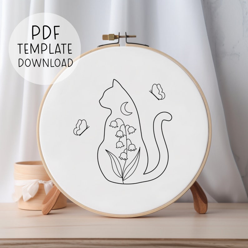 May include: Black and white line art embroidery pattern of a cat sitting with a crescent moon and lily of the valley flowers. Two butterflies are flying around the cat. The pattern is on a white embroidery hoop.